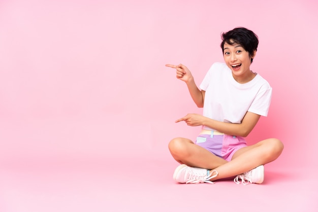 Junge Frau mit kurzen Haaren, die auf dem Boden über vereinzeltem rosa überraschtem und zeigendem Seite sitzen