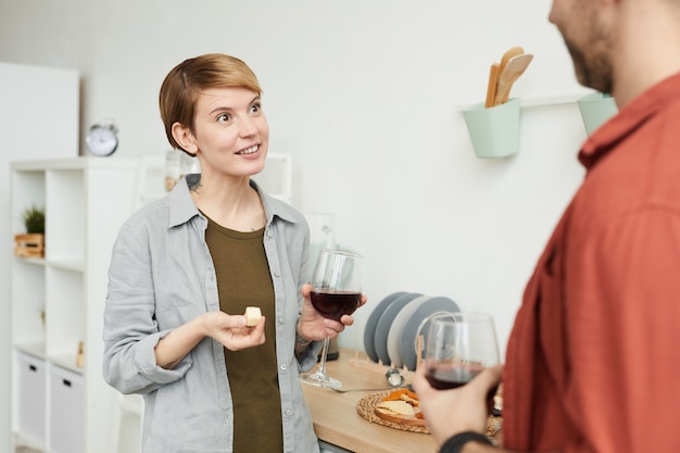 Junge Frau mit kurzen Haaren, die Rotwein trinken und Käse essen und mit Mann sprechen, während sie in der Küche stehen