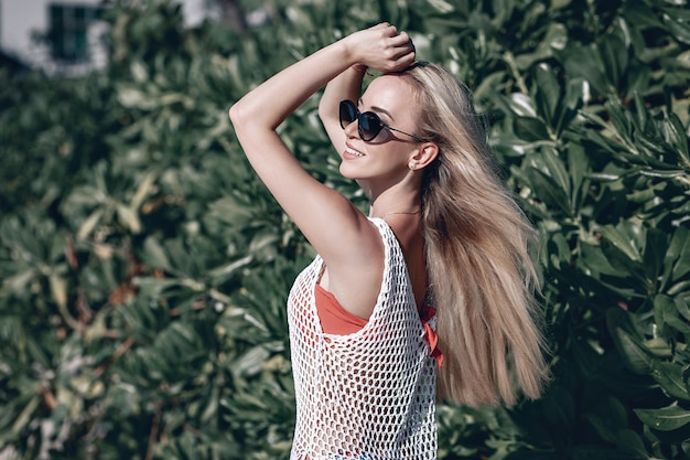 Junge Frau mit langen blonden Haaren und schwarzer Sonnenbrille im süßen weißen Sommerkleid, das in der Nähe von grünen Blättern steht. Mode- und Reisekonzept. Phuket