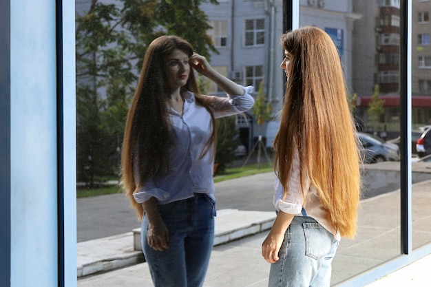 Junge Frau mit langen Haaren betrachtet sich im Spiegelbild des Glasgebäudes