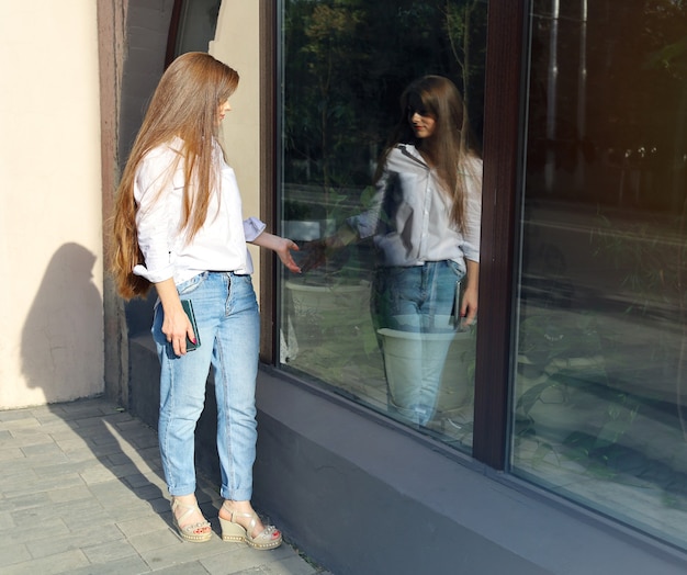 Junge Frau mit langen Haaren steht neben ihrem Spiegelbild in einer Glasvitrine und berührt ihre Hand