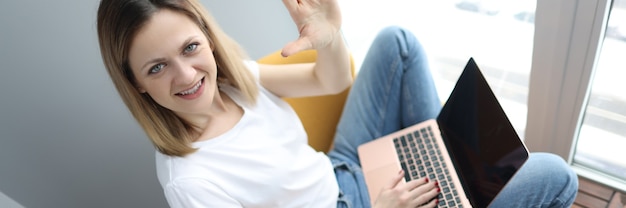 Foto junge frau mit laptop im sessel sitzen und hand winken. telearbeitskonzept