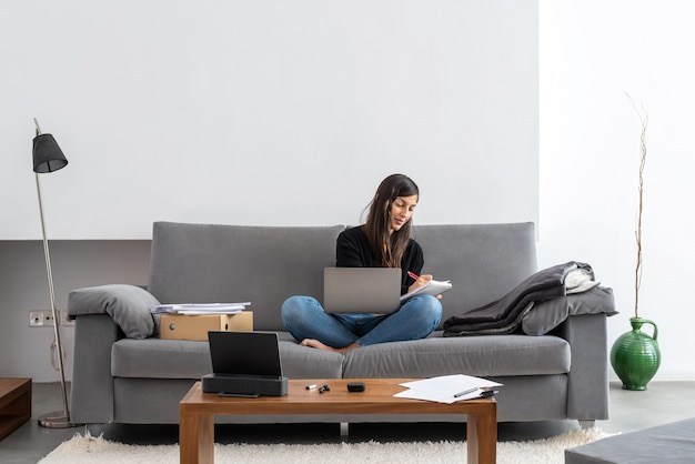 Junge Frau mit Laptop-Telearbeit auf dem Sofa an ihrem Haus