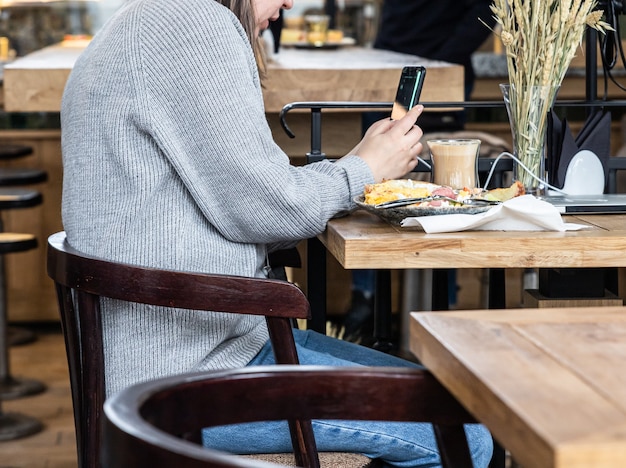 Junge Frau mit leckerem Frühstück mit Omelett, Croissant und Kaffee im Café und mit ihrem Smartphone