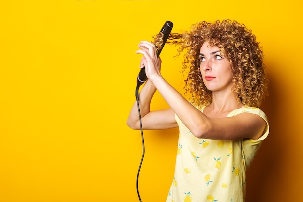 Junge Frau mit lockigem Haar glättet ihr Haar und hält einen Haarglätter auf gelbem Grund.