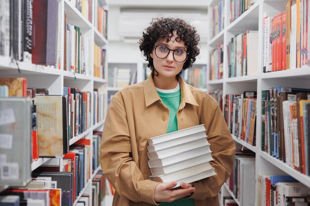 Junge Frau mit lockigem Haar in Bibliothek zwischen Regalen mit Büchern Studentin führt wissenschaftliche Arbeiten durch