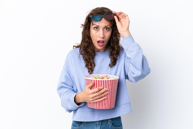 Junge Frau mit lockigem Haar isoliert auf weißem Hintergrund überrascht mit 3D-Brille und hält einen großen Eimer Popcorn