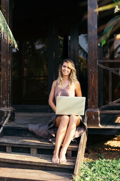 Junge Frau mit losen langen Haaren sitzt barfuß auf Holzstufen der Hütte mit Laptop. Erholung und Tourismus. Heimarbeit. Moderne Technologien. Reisen und Kommunikation. Jugend und Schönheit.