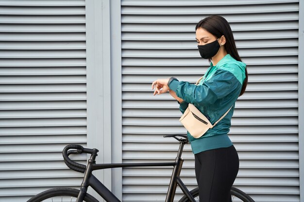 Junge Frau mit Maske, die die Zeit auf der Smartwatch auf der Straße in der Nähe des Fahrrads überprüft. Sport- und Lifestyle-Konzept