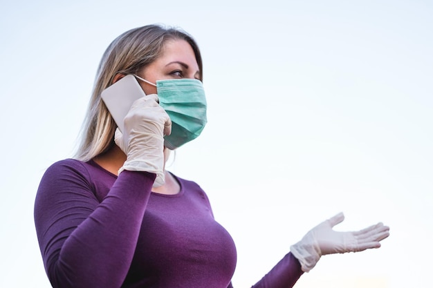 Junge Frau mit Maske spricht über Smartphone vor klarem Himmel