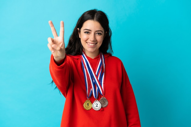Junge Frau mit Medaillen lokalisiert auf weißer Wand lächelnd und Siegeszeichen zeigend