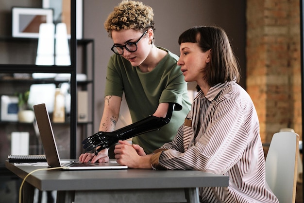 Junge Frau mit prothetischem Arm, die auf Laptop zeigt und während der Arbeit im Büro mit ihrer Kollegin spricht