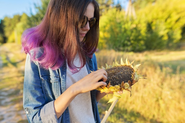 Junge Frau mit reifer Sonnenblumenpflanze in der Hand, schöner Naturlandschaftshintergrund des Sonnenuntergangs. Herbst, Ernte, Landwirtschaft, Gartenarbeit, Sonnenblumenkerne, gesundes natürliches Bio-Lebensmittelkonzept
