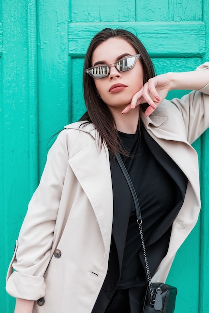 Junge Frau mit Retro-Sonnenbrille im schwarzen Blazer im schwarzen trendigen T-Shirt im stilvollen Frühlings-Trenchcoat steht in der Nähe der hellen Türkistür aus Holz in der Stadt. Städtisches Mädchenmodell im Mode-Outfit.