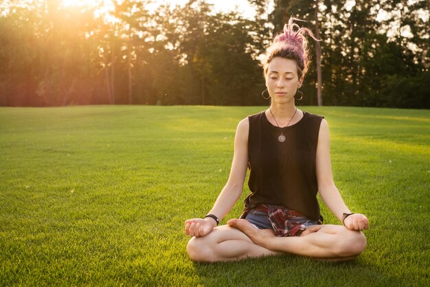 Junge Frau mit rosa Dreadlocks macht Yoga-Übungen im Freien und meditiert im Park bei Sonnenuntergang. Lotussitz