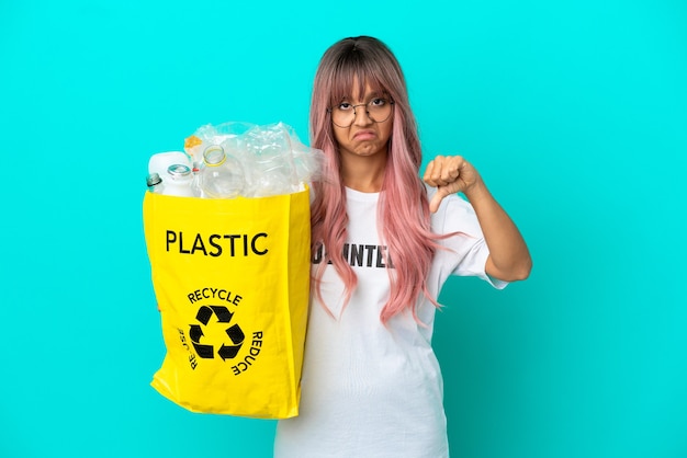 Junge Frau mit rosa Haaren, die eine Tasche voller Plastikflaschen zum Recyceln hält, isoliert auf blauem Hintergrund, der Daumen nach unten mit negativem Ausdruck zeigt