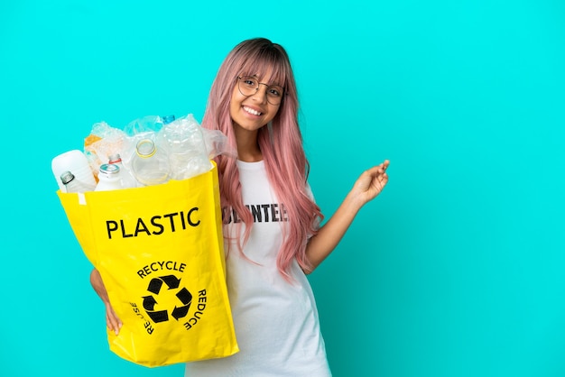 Junge Frau mit rosa Haaren, die eine Tasche voller Plastikflaschen zum Recyceln hält, isoliert auf blauem Hintergrund, der nach hinten zeigt