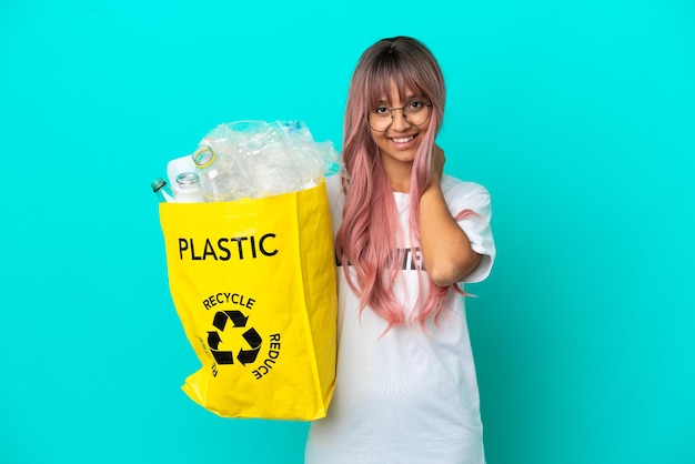 Junge Frau mit rosa Haaren, die eine Tasche voller Plastikflaschen zum Recyceln hält, isoliert auf blauem Hintergrund lachend