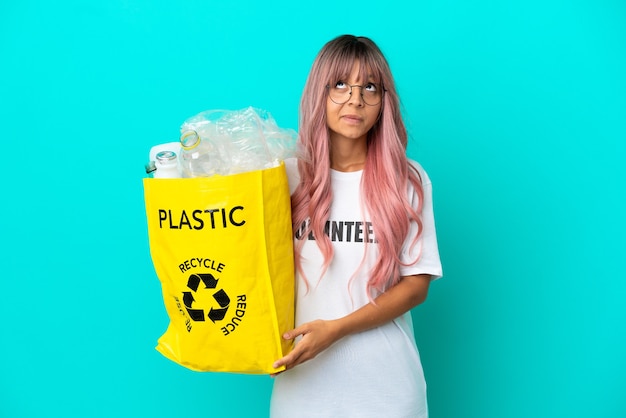Junge Frau mit rosa Haaren, die eine Tasche voller Plastikflaschen zum Recyceln hält, isoliert auf blauem Hintergrund und nach oben schauend