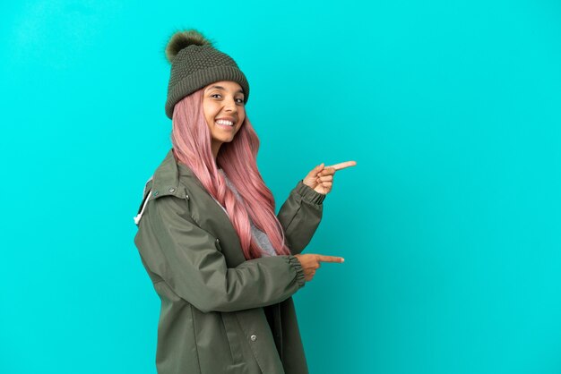 Junge Frau mit rosa Haaren, die einen regendichten Mantel trägt, isoliert auf blauem Hintergrund, der mit dem Finger zur Seite zeigt und ein Produkt präsentiert