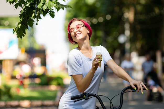 Junge Frau mit rosa Haaren geht in den Park mit einem Fahrrad, das im Sommer Eis isst. Umwelttransportmittel