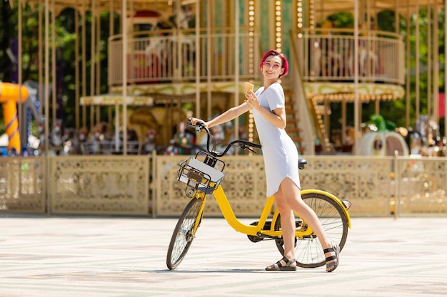 Junge Frau mit rosa Haaren geht in den Park mit einem Fahrrad, das im Sommer Eis isst.