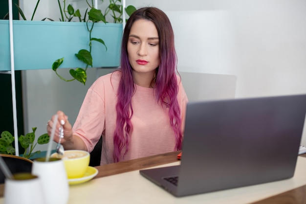 Junge Frau mit rosa Haaren mit Laptop, die im Café sitzt, intelligente Studentin, die nach ihren Vorlesungen an der Universität an einem Netbook arbeitet