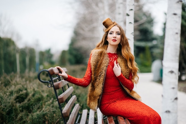 Junge Frau mit roten Haaren in einem leuchtend roten Kleid auf einer Bank in einem leeren Park unter den Birkenbir