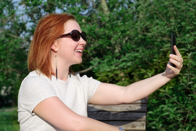Junge Frau mit roten Haaren in Sonnenbrille lächelt mit ausgestreckter Hand auf den Bildschirm seines Smartphones, um ein Foto von sich selbst am Telefon zu machen. Frau macht ein Selfie auf einem Smartphone in einem Stadtpark.