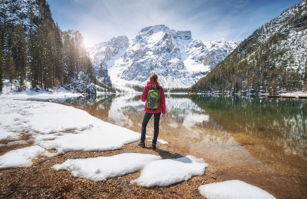 Junge Frau mit Rucksack am verschneiten Ufer des Pragser Wildsees mit klarem Wasser an einem sonnigen, hellen Tag im Frühling. Reiselandschaft mit schlankem Mädchen, das sich im Wasser spiegelt, Berge, grüne Bäume, blauer Himmel