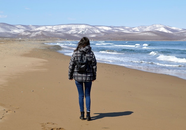 Junge Frau mit Rucksack, der auf dem Strand nahe den schneebedeckten Bergen und dem japanischen Meer geht