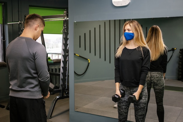 Junge Frau mit Schutzmaske, die mit Personal Trainer im Fitnessstudio während der Covid-19-Pandemie trainiert. Sie pumpt ihre Muskulatur mit einer Hantel auf.