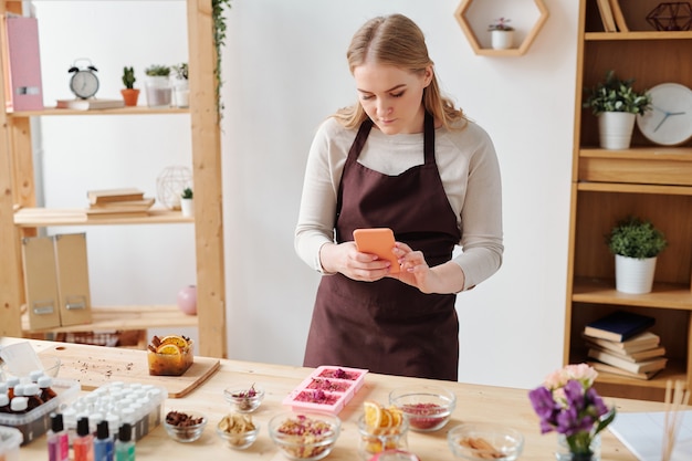 Junge Frau mit Smartphone, die Foto von handgemachter Seife in Silikonformen auf Holztisch im Studio macht