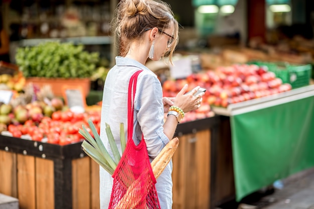 Junge Frau mit Smartphone steht mit Tasche vor dem Lebensmittelmarkt in Frankreich