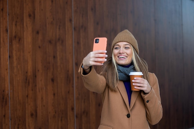 Junge Frau mit Smartphone vor verschwommenem Gebäudehintergrund auf der Straße Modefoto eines schönen Mädchens mit Telefon und einer Tasse Kaffee