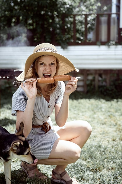 Foto junge frau mit sommerhut grillt fleisch im freien im hinterhof, sitzt mit ihrem hund und gibt haustier einen snack