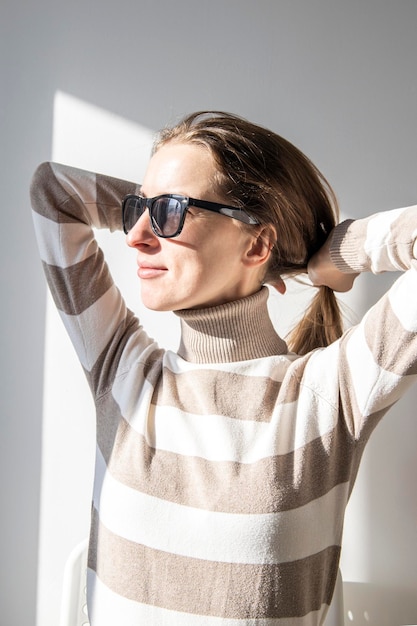 Junge Frau mit Sonnenbrille glättet ihr Haar an einer weißen Wand
