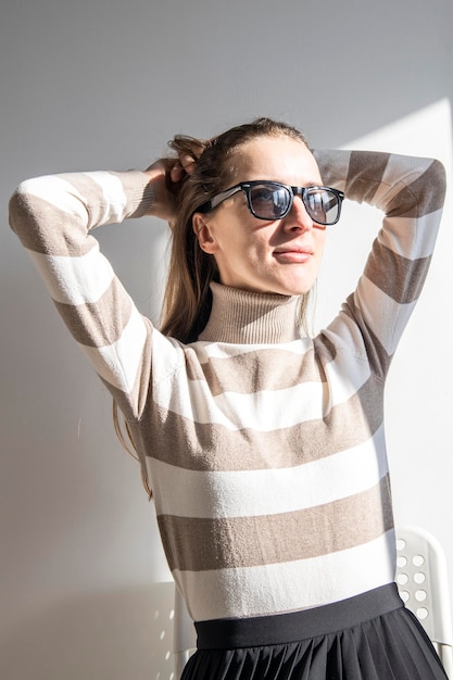 Junge Frau mit Sonnenbrille in einem gestreiften Pullover vor einer weißen Wand