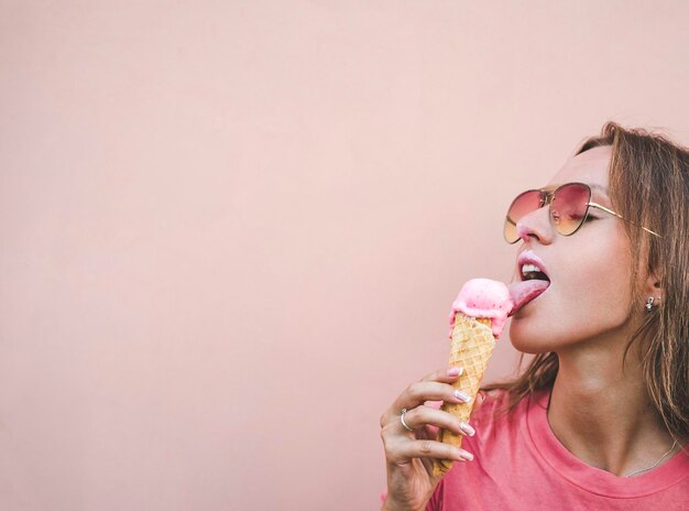 Junge Frau mit Sonnenbrille leckt geschmolzenes rosa Erdbeereis auf rosa Hintergrund Textfreiraum