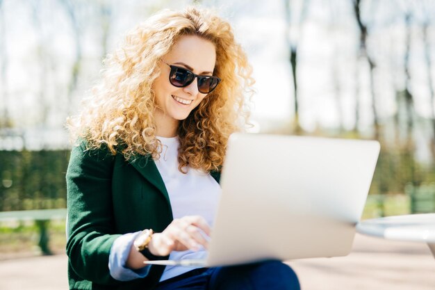 Foto junge frau mit sonnenbrille mit flauschigem haar, die fröhlich lächelt und sich im park ausruht, indem sie mit einem laptop-computer im internet surft und in sozialen netzwerken surft menschen-lifestyle-technologie und erholungskonzept