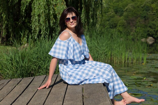 Junge Frau mit Sonnenbrille sitzt an einem sonnigen Tag auf der Brücke in der Nähe des Flusses