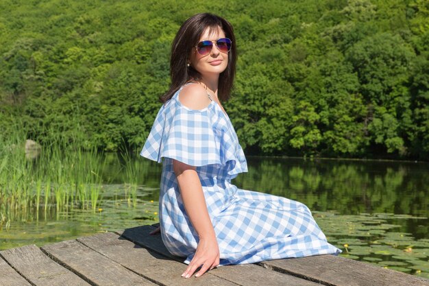 Junge Frau mit Sonnenbrille sitzt an einem sonnigen Tag auf der Brücke in der Nähe des Flusses