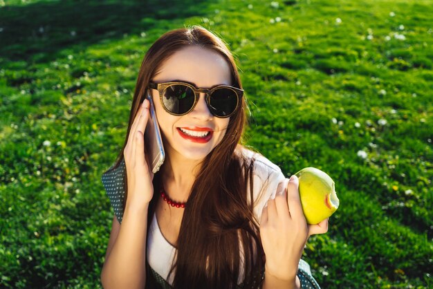 Junge Frau mit Sonnenbrille sitzt im Park, auf dem Rasen mit hellgrünem Gras, lächelt, isst einen Apfel, telefoniert