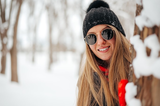 Junge Frau mit Sonnenbrille und Hut versteckt sich hinter dem Baum in einem verschneiten Park
