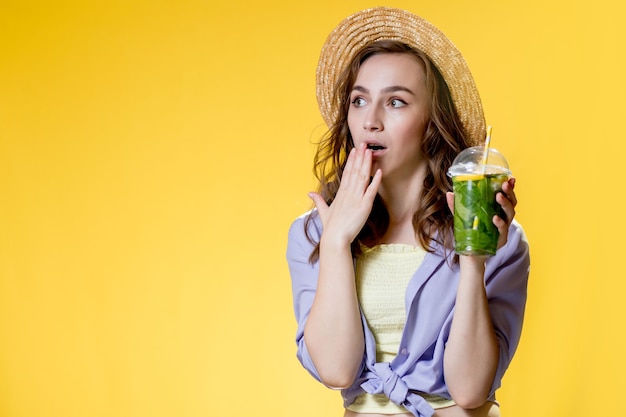 Junge Frau mit Strohhut, die ein Plastikbechergetränk hält und auf gelbem Hintergrund überrascht aussieht