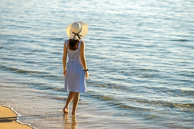 Junge Frau mit Strohhut und einem Kleid, das allein am leeren Sandstrand am Meeresufer steht. Einsames touristisches Mädchen, das auf Urlaubsreise den Horizont über der ruhigen Meeresoberfläche betrachtet.