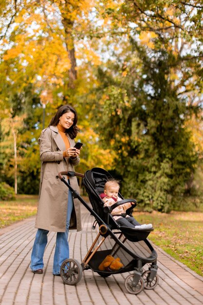 Foto junge frau mit süßem baby in einem kinderwagen benutzt im herbstpark ein handy