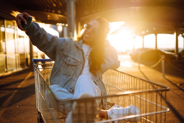 Junge Frau mit Telefon sitzt Einkaufswagen und hat Spaß in der Nähe der Farbwand Blogging Shopaholic