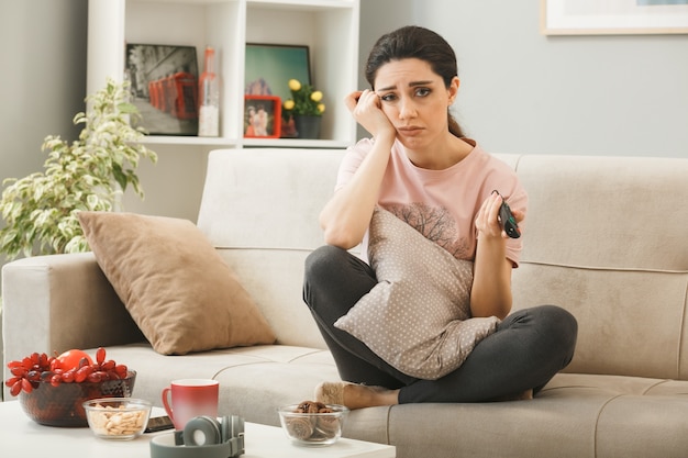 Junge Frau mit TV-Fernbedienung sitzt auf dem Sofa hinter dem Couchtisch im Wohnzimmer