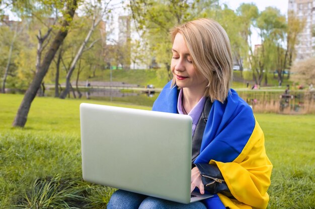 Junge Frau mit ukrainischer Flagge, die am Laptop im Stadtpark arbeitet Fernarbeit Freier Lebensstil